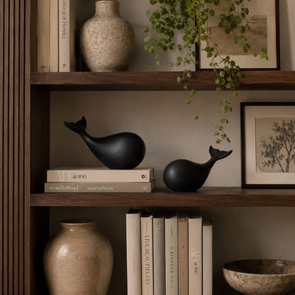 Decorative black whale sculpture set displayed on a wooden bookshelf with books, ceramic vase, and indoor plants.
