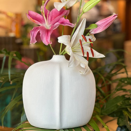 A handcrafted white ceramic vase with a unique vertical seam and textured finish, featuring fresh pink and white lilies in a warm, elegantly lit room.