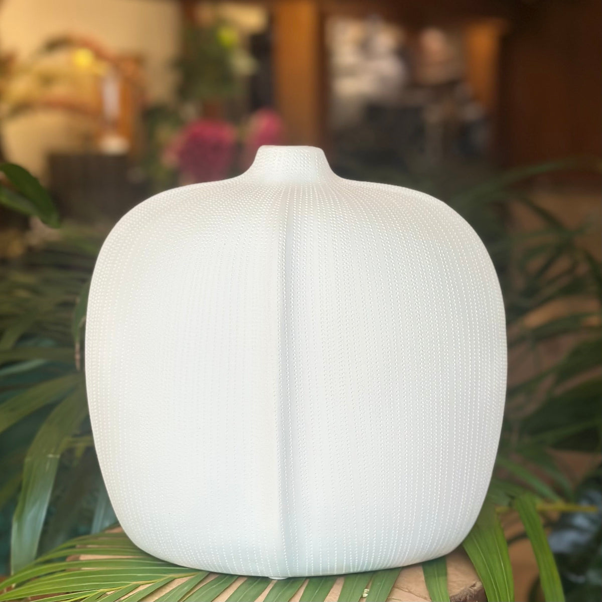 A close-up of a premium white ceramic bud vase with a fine dotted texture and a minimalist rounded silhouette, displayed on a natural wood block with tropical greenery in the background.