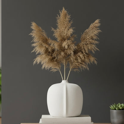 A modern white textured vase holding dried pampas grass, styled on a stack of books against a dark grey wall for a contemporary luxury home decor aesthetic.