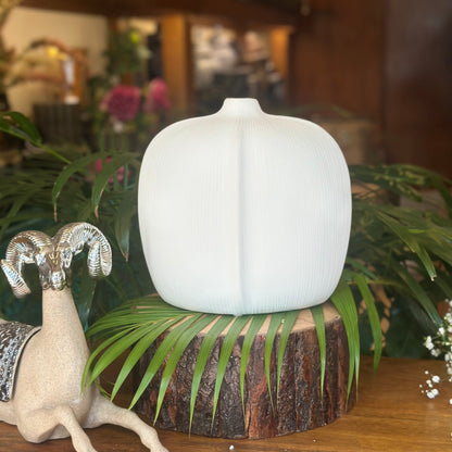 A modern, matte white ceramic bud vase with a fine ribbed texture, shaped like a stylized pumpkin, displayed on a wooden log base among green tropical leaves