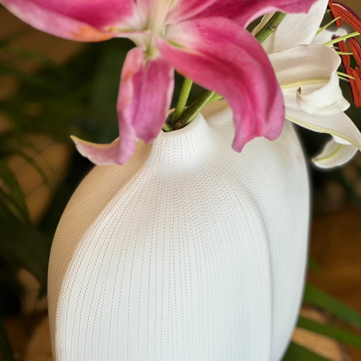 A close-up view of a white ceramic vase featuring a fine, dotted ribbed texture and a narrow neck, holding a vibrant pink lily and a white lily.