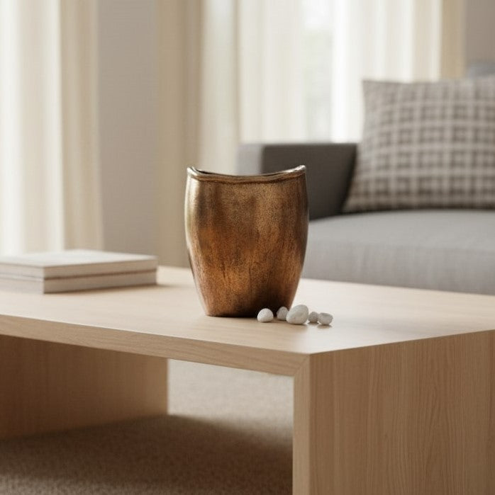 An empty, oval-shaped bronze vase with a rustic textured finish sitting on a light oak coffee table next to small white decorative stones.