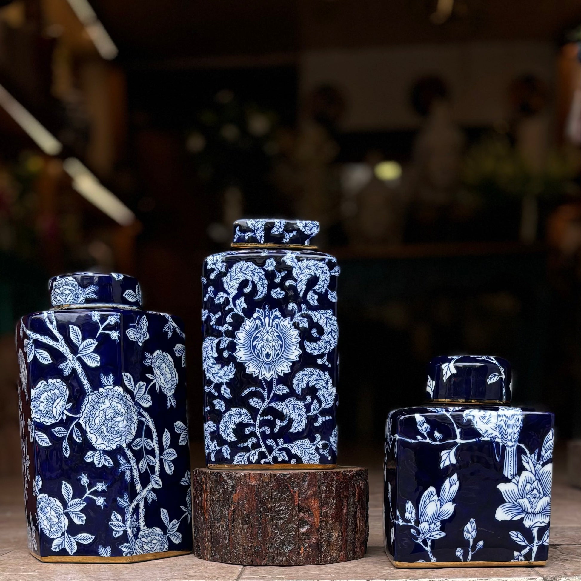 A set of three graduated navy blue and white porcelain ginger jars with intricate floral and botanical patterns, featuring elegant gold-rimmed bases and lids, displayed on a rustic wood pedestal.