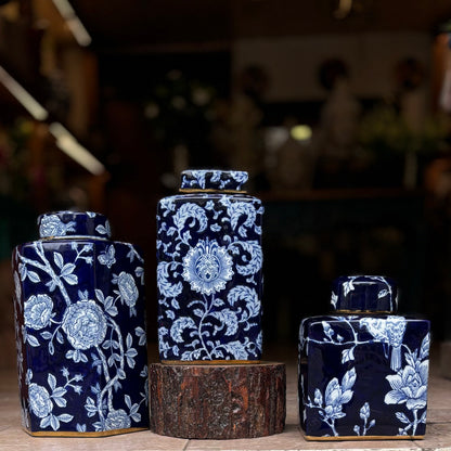 A set of three graduated navy blue and white porcelain ginger jars with intricate floral and botanical patterns, featuring elegant gold-rimmed bases and lids, displayed on a rustic wood pedestal.