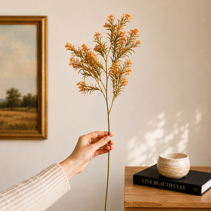 Hand holding a single orange artificial floral stem in a warm, modern home setting with neutral decor, wooden console table, and framed artwork in the background.
