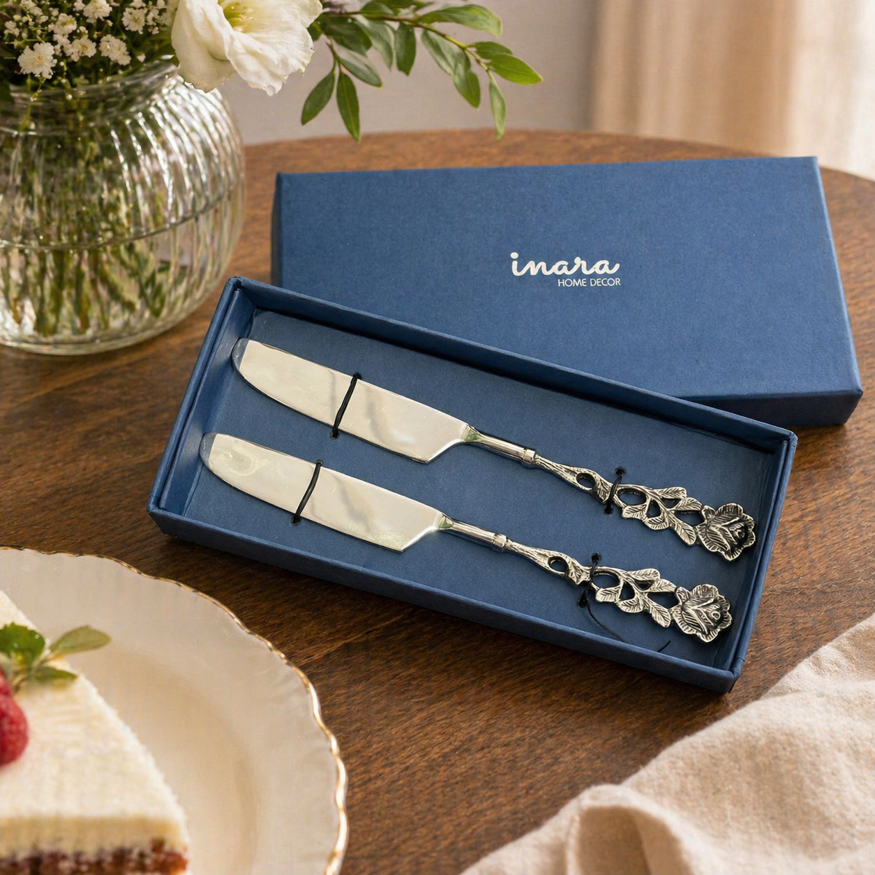 Ornate silver cake server and knife set with floral handles displayed in a blue gift box on a wooden table with cake and flowers in a lifestyle setting.