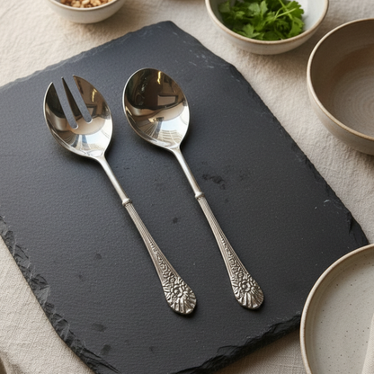 Flatlay view of a pair of heavy silver salad servers on a black slate stone, highlighting the decorative embossed floral patterns on the handles.