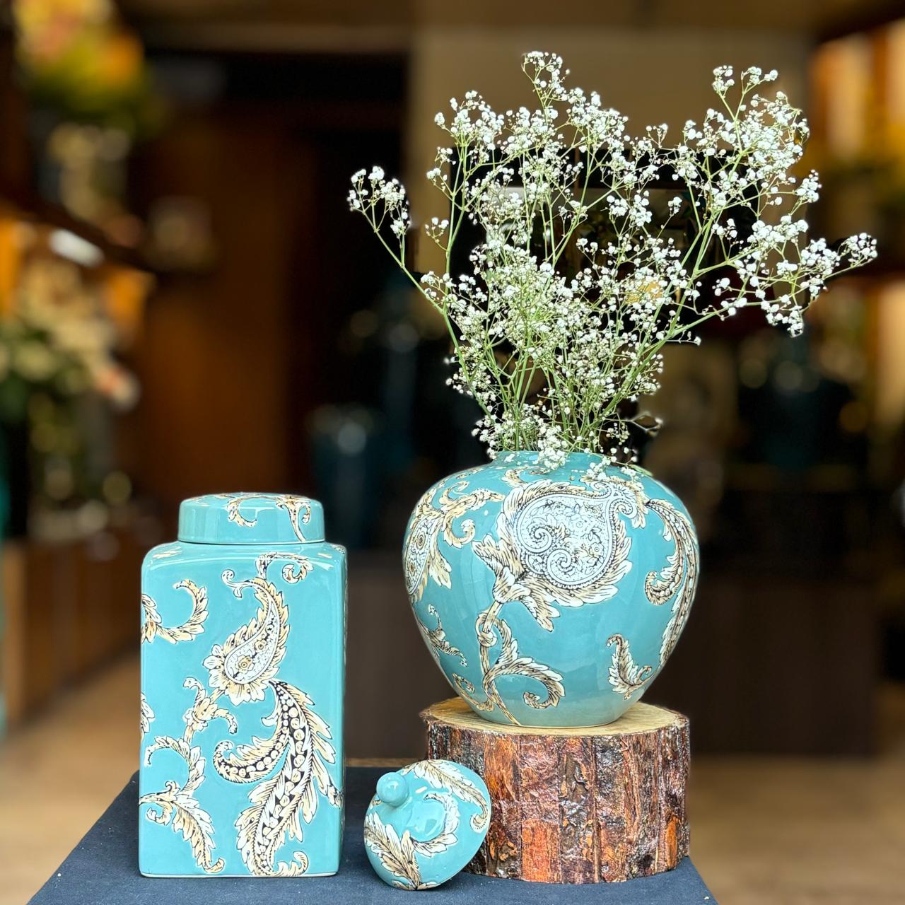 A premium lifestyle arrangement of turquoise paisley ceramic jars; the round jar is used as a vase holding delicate white flowers, set against a warm, blurred interior background.