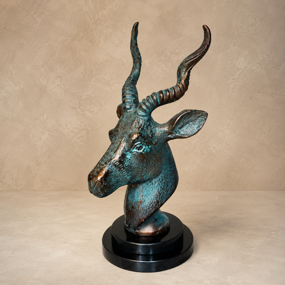 Patina antelope head sculpture with verdigris finish and black circular base displayed on a luxury beige studio backdrop, premium decorative accent for modern home décor.