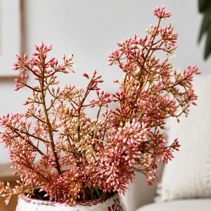 Close-up lifestyle image of a pink artificial floral arrangement displayed in a decorative white and red ceramic vase on a wooden coffee table in a modern living room setting.