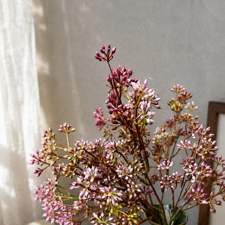 Close-up lifestyle image of pink artificial wildflower stems arranged in a glossy green ceramic vase, styled on a wooden table with soft natural sunlight and a neutral textured wall background.