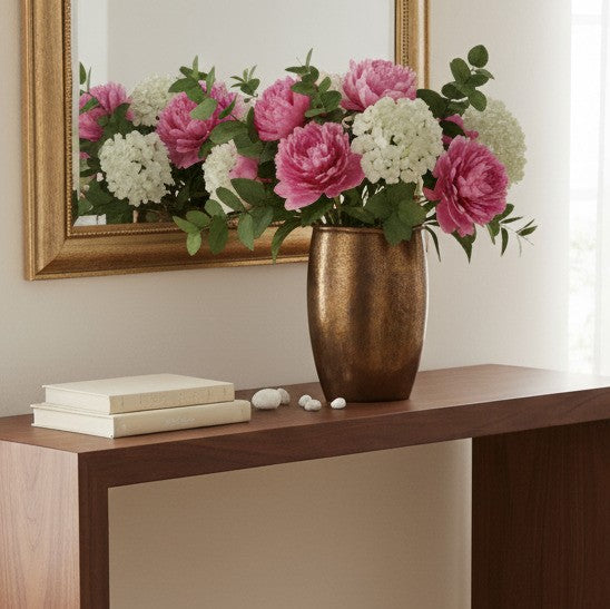 A lush floral arrangement of bright pink peonies and white hydrangeas with eucalyptus leaves in a textured bronze vase, sitting on a dark wood console table in front of a gold-framed mirror.
