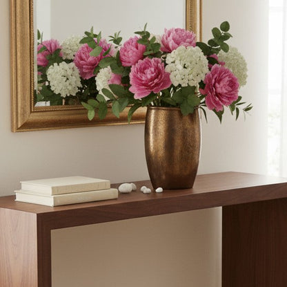 A lush floral arrangement of bright pink peonies and white hydrangeas with eucalyptus leaves in a textured bronze vase, sitting on a dark wood console table in front of a gold-framed mirror.