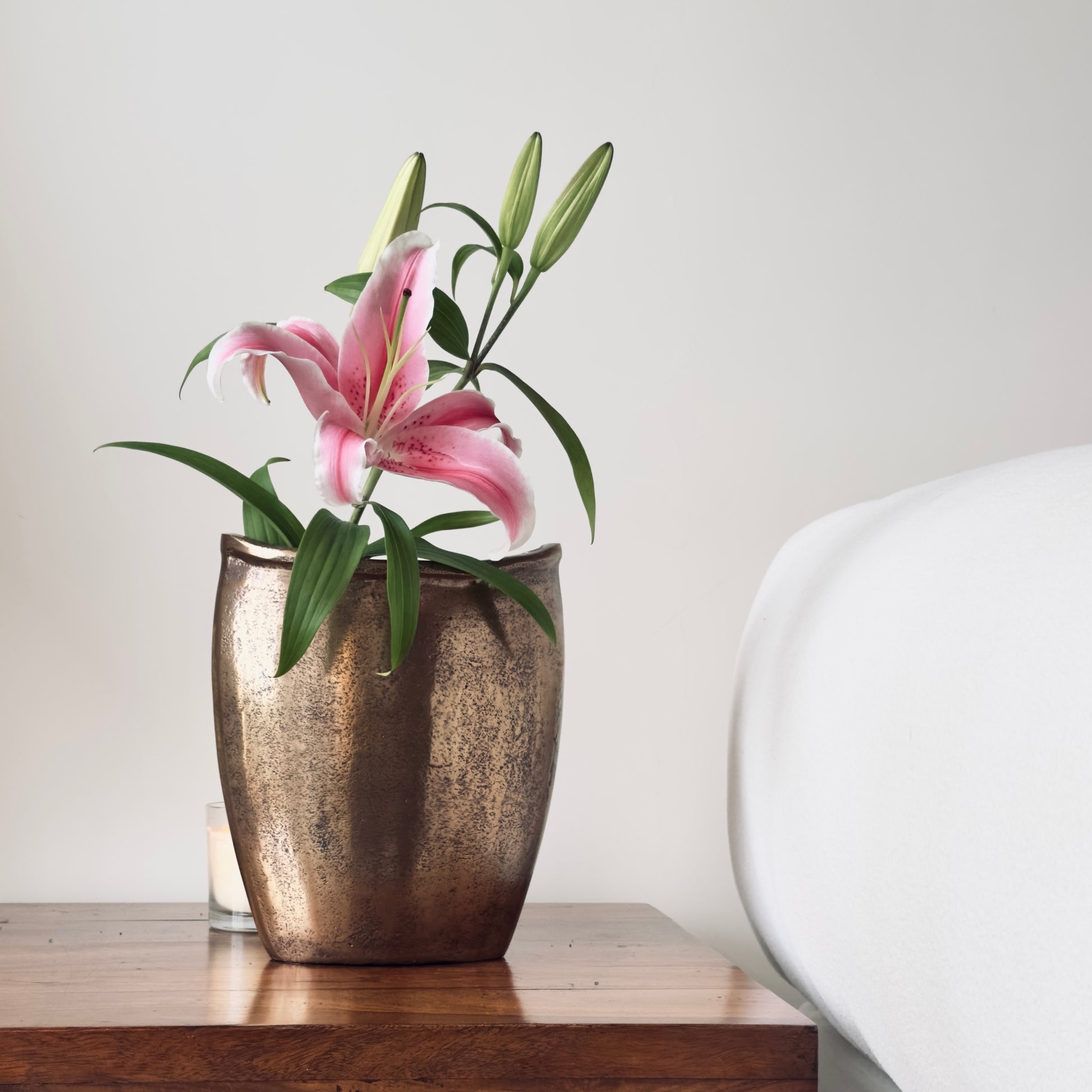 A single pink Stargazer lily with green buds in a rustic bronze vase, placed on a dark wood bedside table against a clean white wall.