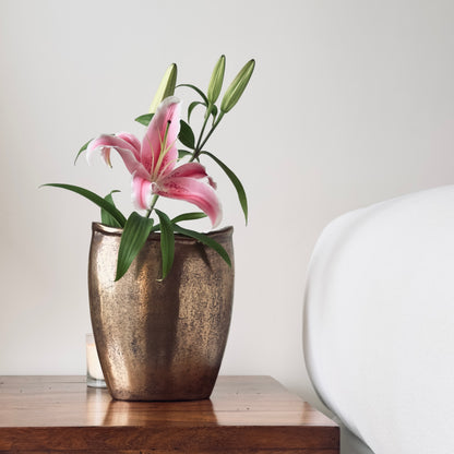 A single pink Stargazer lily with green buds in a rustic bronze vase, placed on a dark wood bedside table against a clean white wall.