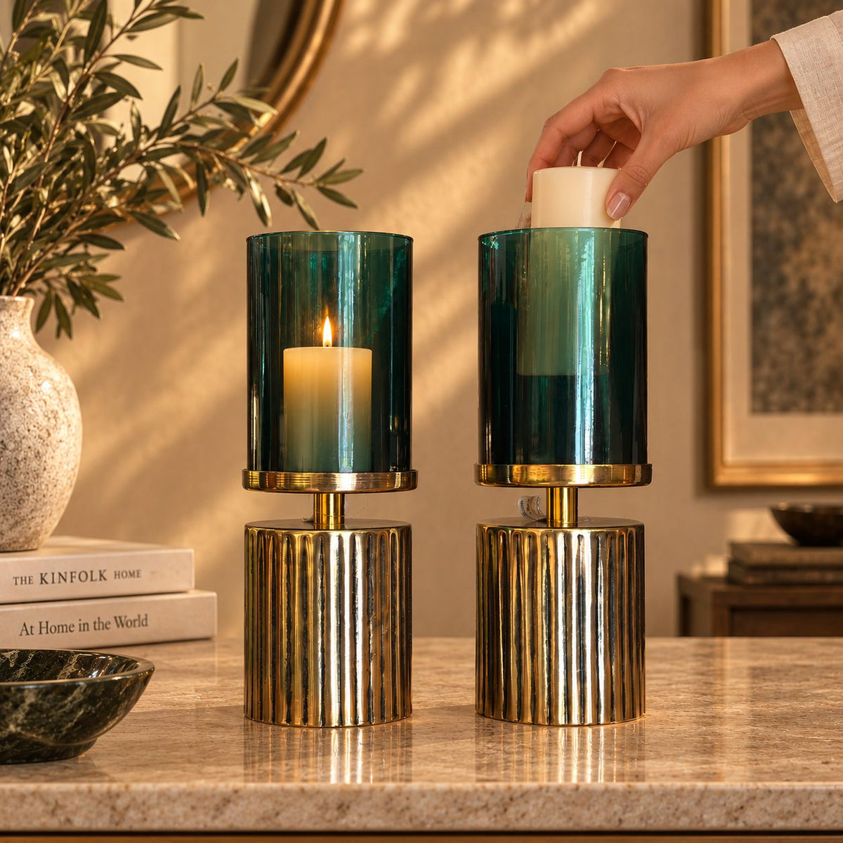 Hand placing a cream pillar candle into a green glass hurricane candle holder while the second holder is lit, demonstrating product use.