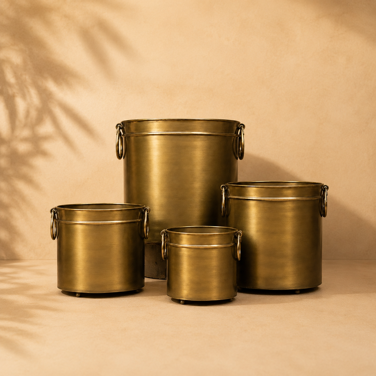 Antique brass indoor planter set featuring four luxury metal planters in assorted sizes with side ring handles, displayed against a warm beige Venetian plaster wall and matte microcement floor in a premium studio setting.