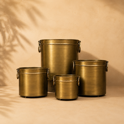 Antique brass indoor planter set featuring four luxury metal planters in assorted sizes with side ring handles, displayed against a warm beige Venetian plaster wall and matte microcement floor in a premium studio setting.