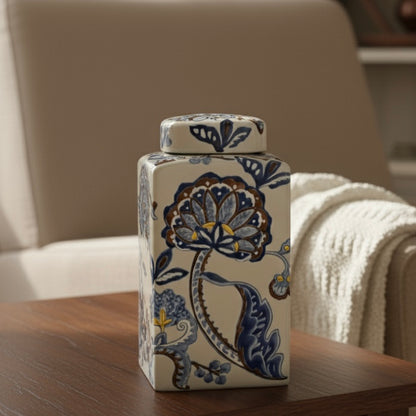 Premium rectangular ceramic storage jar featuring intricate ikat floral artwork in cobalt blue, placed on a dark wood side table with a cream-colored throw blanket and soft armchair in the blurred background.