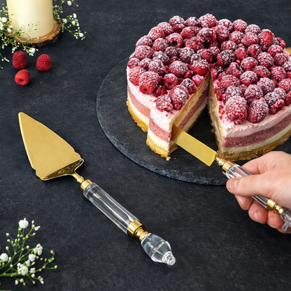 Premium gold cake server and knife set with crystal handles cutting raspberry layer cake on black stone plate, elegant wedding cake servi