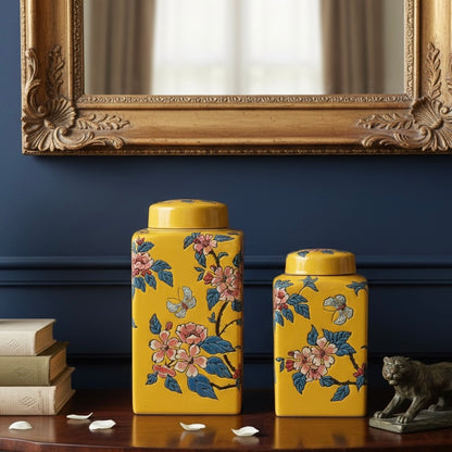 Pair of decorative yellow floral jars placed on a dark wood sideboard in front of a navy blue wall and ornate gold mirror.