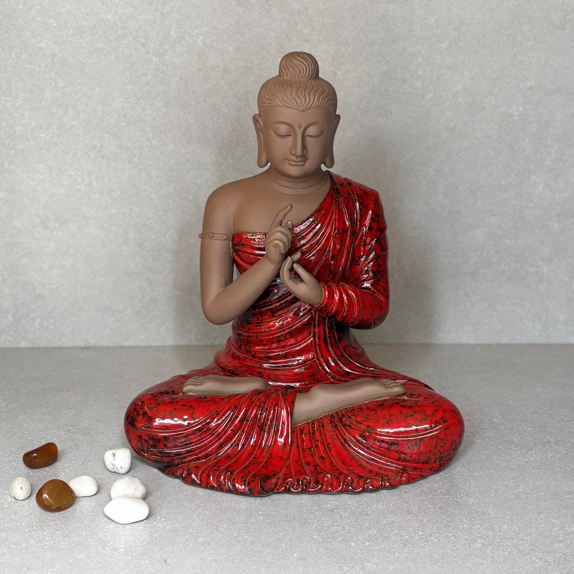 A handcrafted ceramic Buddha statue with a textured red robe and matte brown skin, sitting in a meditative pose on a neutral background with small river stones.