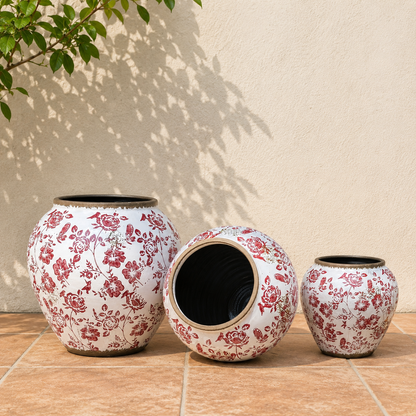 Lifestyle image of red floral ceramic planters with vintage botanical patterns placed against a neutral wall in a sunlit outdoor setting.