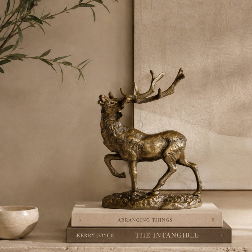 Luxury bronze stag decorative sculpture styled on stacked coffee table books atop a travertine console, with olive branches and a warm beige textured wall in a premium contemporary home interior.