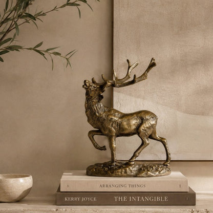 Luxury bronze stag decorative sculpture styled on stacked coffee table books atop a travertine console, with olive branches and a warm beige textured wall in a premium contemporary home interior.