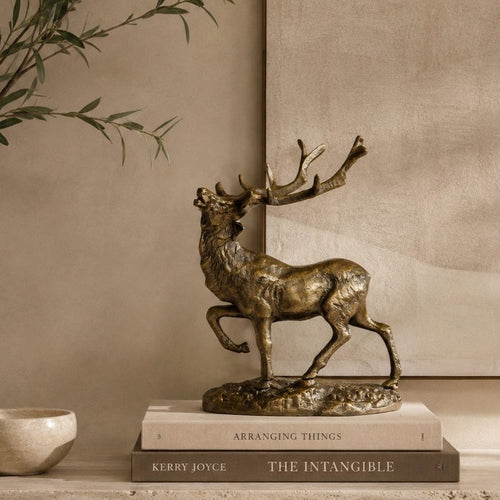 Luxury bronze stag decorative sculpture styled on stacked coffee table books atop a travertine console, with olive branches and a warm beige textured wall in a premium contemporary home interior.