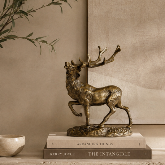Luxury bronze stag decorative sculpture styled on stacked coffee table books atop a travertine console, with olive branches and a warm beige textured wall in a premium contemporary home interior.