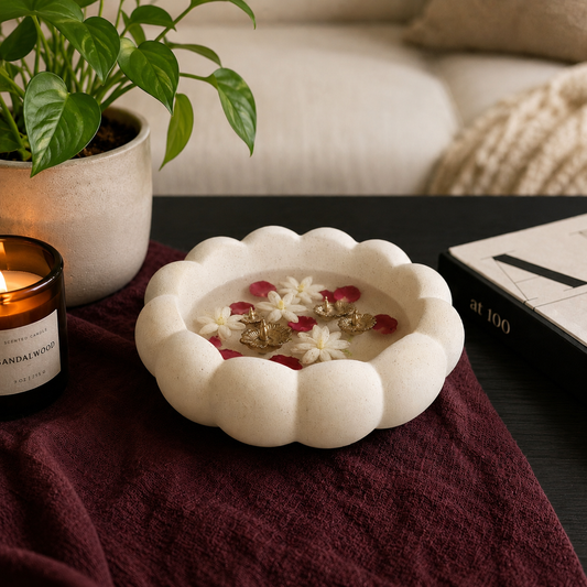 White scalloped ceramic urli with floating floral trinkets styled on a dark black table with dark maroon fabric, candle, and indoor plant decor.