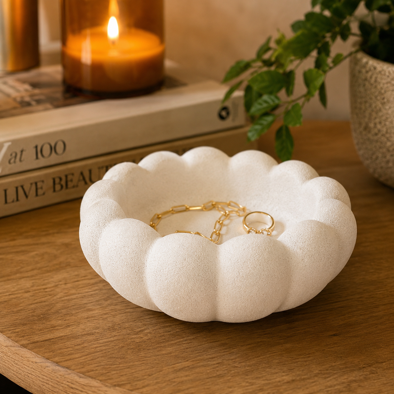Close-up lifestyle image of a cream textured scalloped decorative bowl styled with gold jewelry on a wooden table with warm candlelight and modern decor accents.