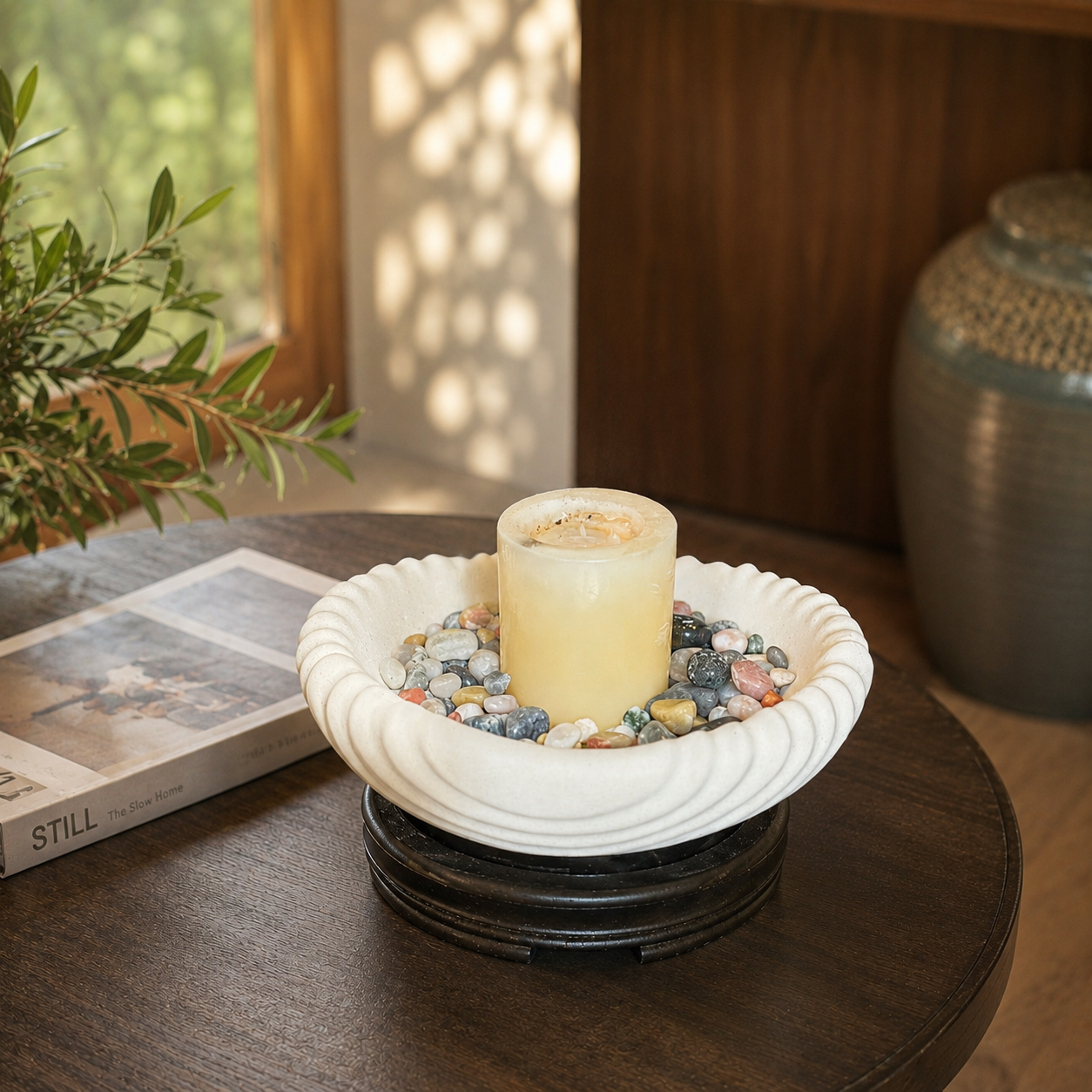 Scalloped white ceramic decorative urli with colorful pebbles and pillar candle styled on a wooden coffee table in a modern living room setting.