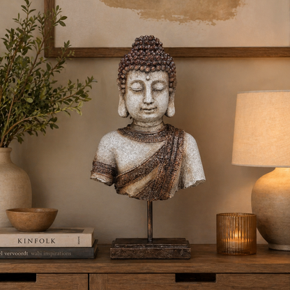 Antique finish Buddha bust sculpture displayed on a wooden console table with warm ambient lighting, decorative books, candle holder, and indoor plant in a serene modern interior setup.