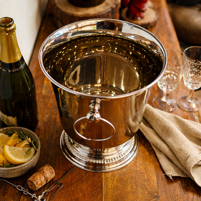 Polished silver champagne bucket styled on a wooden table with champagne bottle, crystal glasses, lemon slices, and linen napkin in an elegant lifestyle setting.