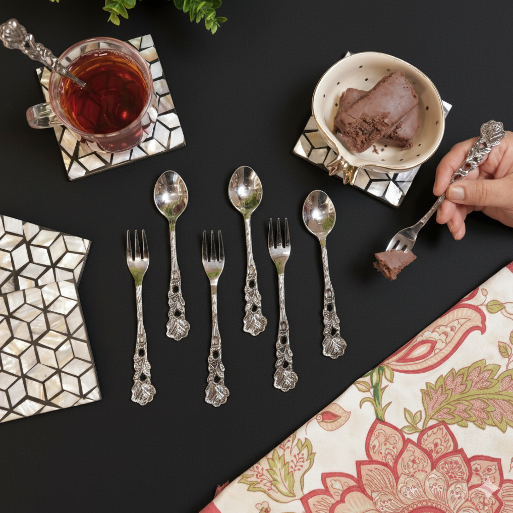 Six embossed silver spoons arranged on a black tabletop next to a white sugar bowl, cream pitcher, and a glass of dark tea on a geometric inlay coaster.