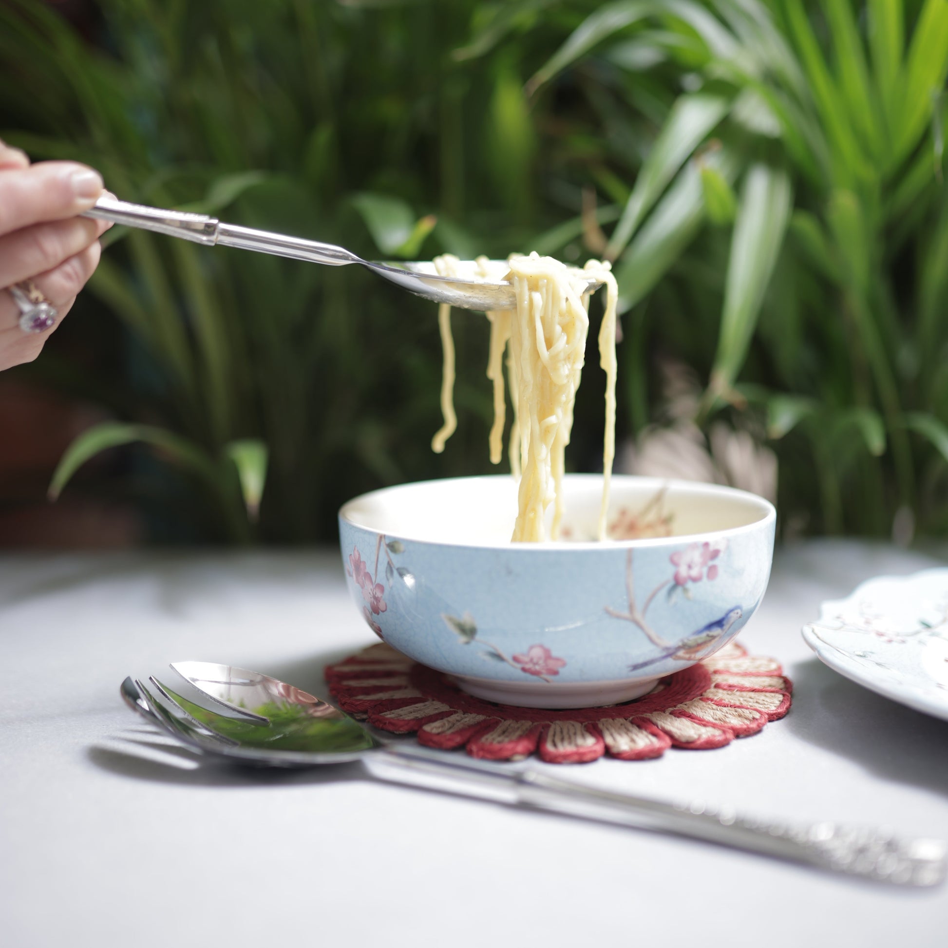 Close-up of a silver serving spoon lifting a portion of noodles from a blue floral bowl, showing the intricate floral detailing on the handle.