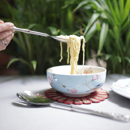 Close-up of a silver serving spoon lifting a portion of noodles from a blue floral bowl, showing the intricate floral detailing on the handle.