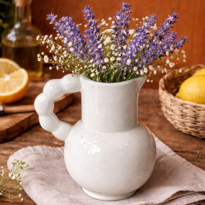 White ceramic loop-handle vase filled with lavender and baby’s breath flowers, styled on a wooden kitchen table with lemons and warm rustic décor in the background.