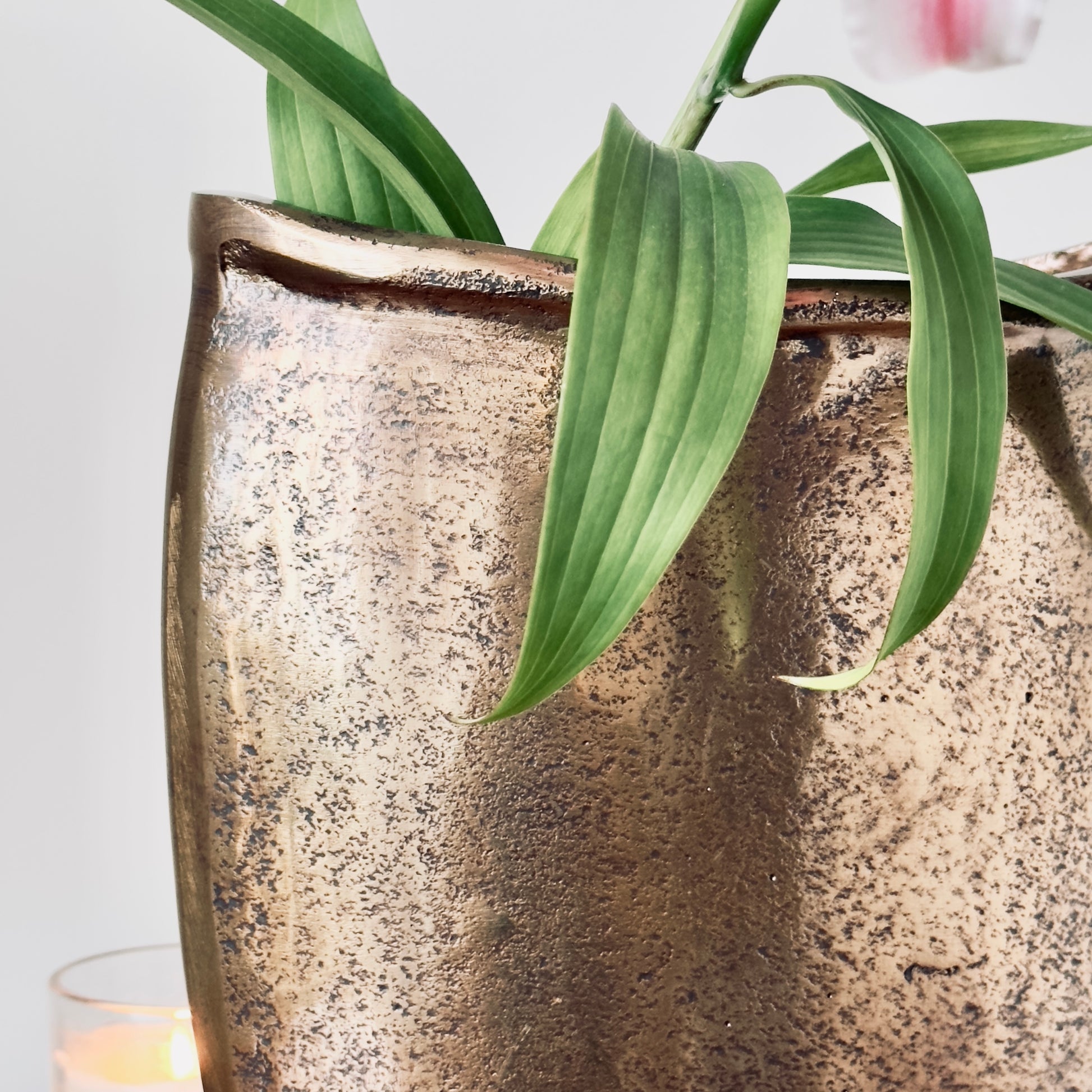A detailed close-up shot showing the pitted, metallic texture of a bronze vase with vibrant green lily leaves emerging from the top.