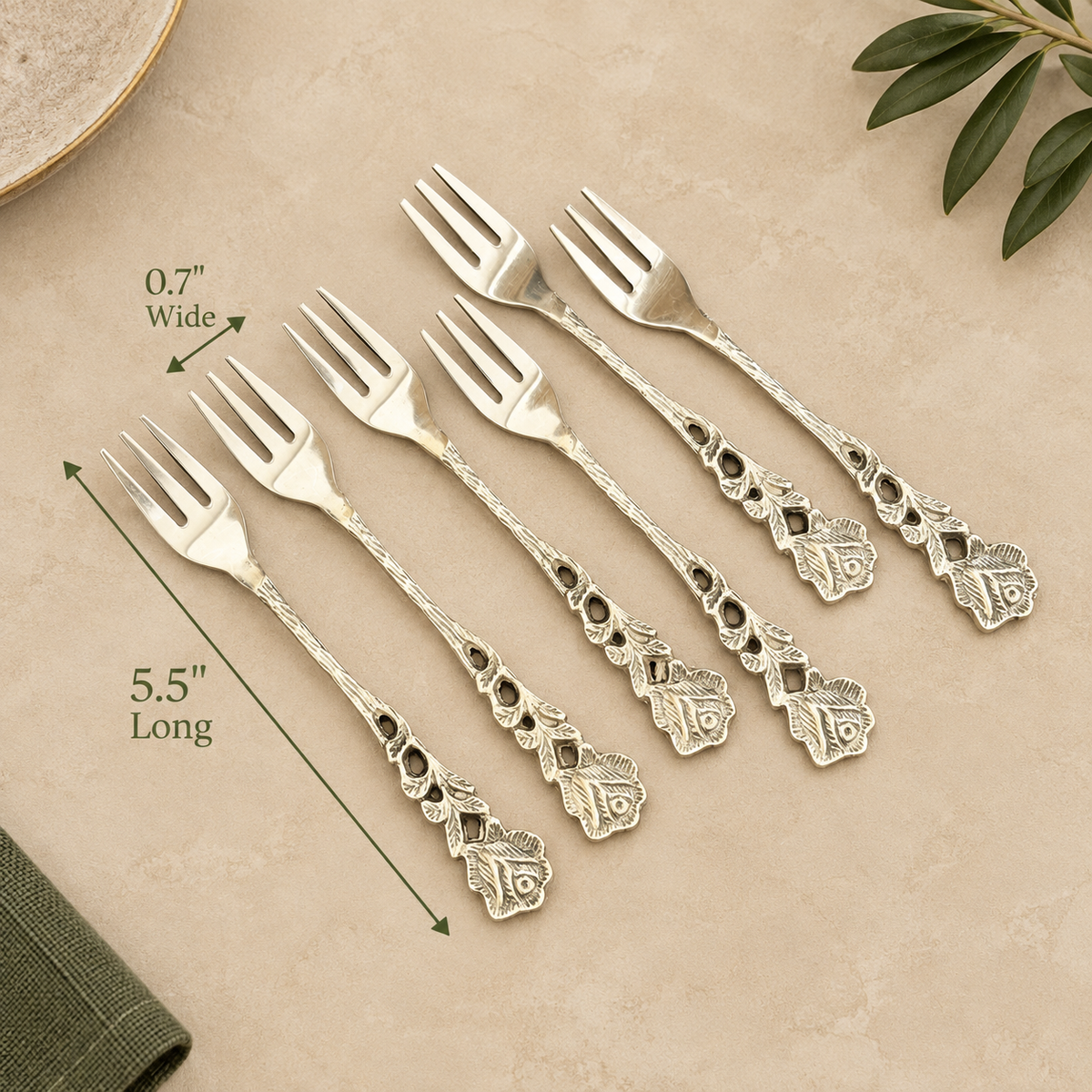 Vintage antique silver dessert forks set of 6 with ornate floral handles displayed on a warm beige Venetian seamless matte microcement surface, featuring elegant olive green dimension arrows showing 5.5 inches long and 0.7 inches wide.