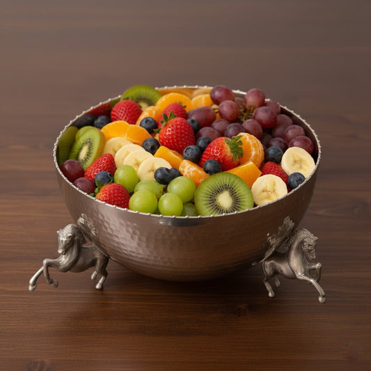 Vintage hammered metal fruit bowl with decorative horse legs, filled with fresh fruits, styled on a wooden dining table with flowers and drinks in the background.