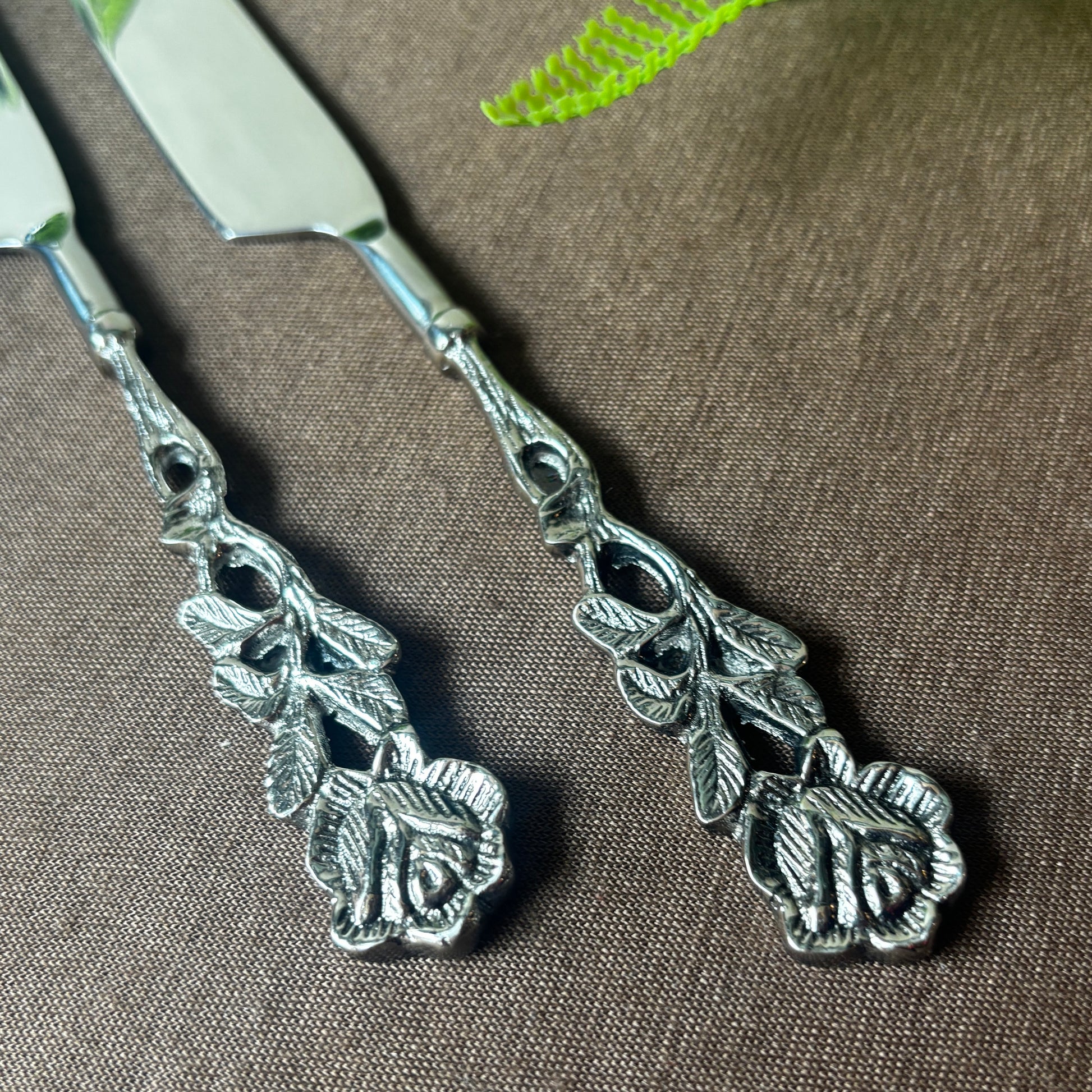 Detailed close-up of two silver-toned butter knives featuring intricately carved rose and leaf handles on a textured brown background.