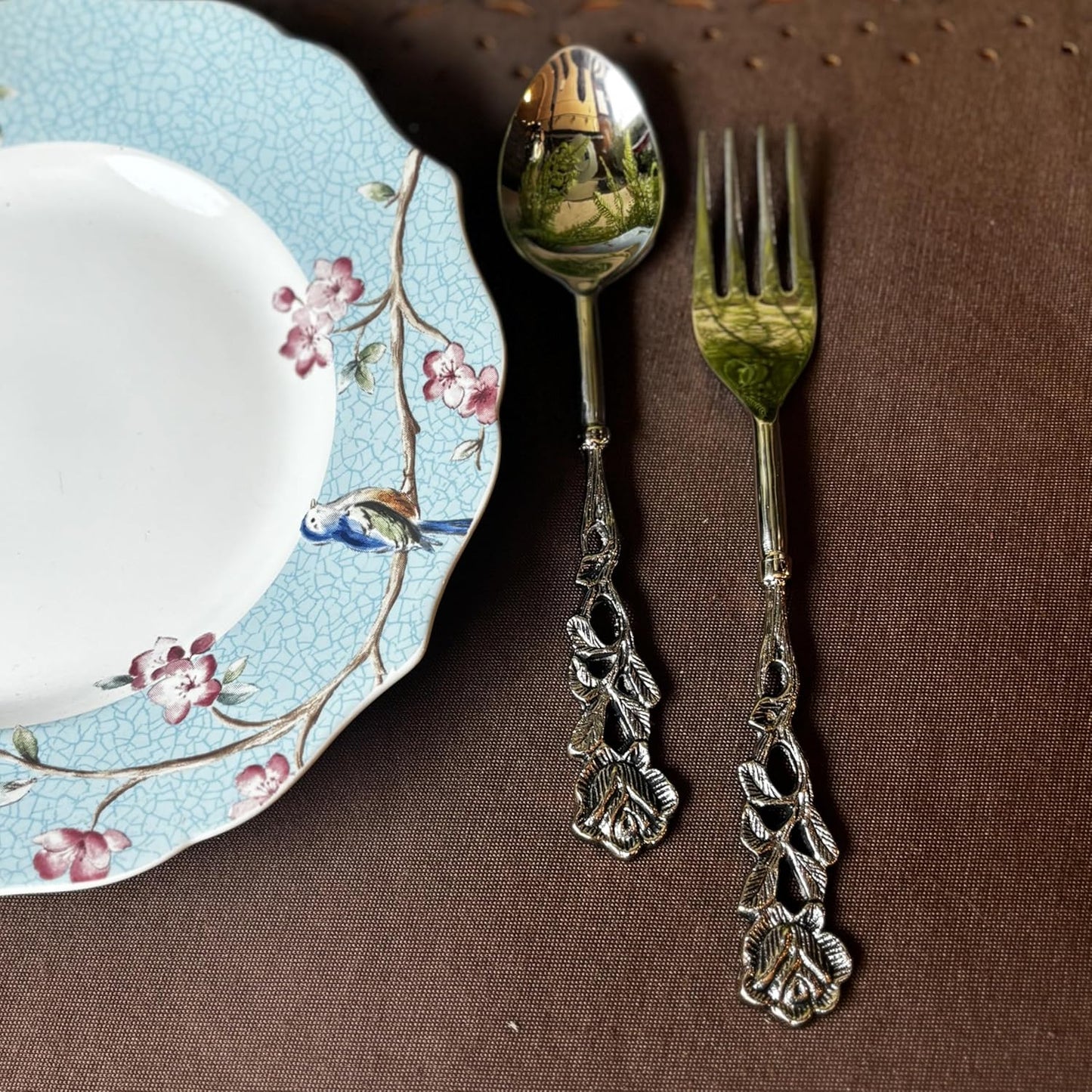 Close-up of a silver-finished dessert spoon and fork with rose-patterned handles next to a light blue floral ceramic plate.