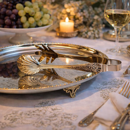 Elegant gold-tone shell-shaped serving tongs resting on an ornate silver footed tray, set on a formal dining table with candlelight and grapes.