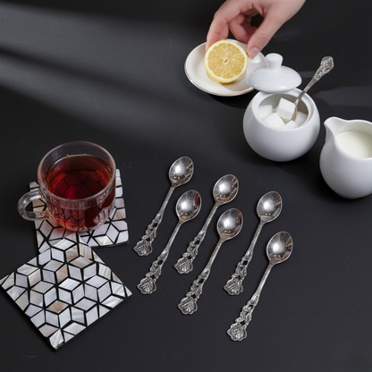 Six embossed silver spoons arranged on a black tabletop next to a white sugar bowl, cream pitcher, and a glass of dark tea on a geometric inlay coaster.