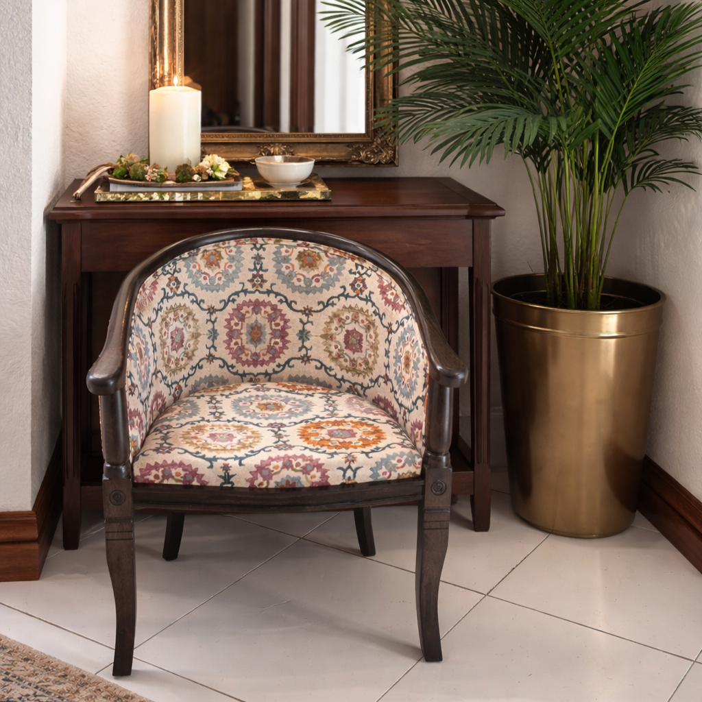 Upholstered patterned barrel accent chair styled in front of a dark wood console table with an antique framed mirror, decorative tray with candle and bowl, and a tall statement plant in a brass planter.