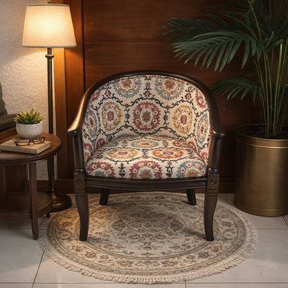 Patterned upholstered barrel accent chair with dark wooden frame styled beside a round wooden side table with lamp and books, placed on a round rug with a tall palm plant in a brass planter behind.